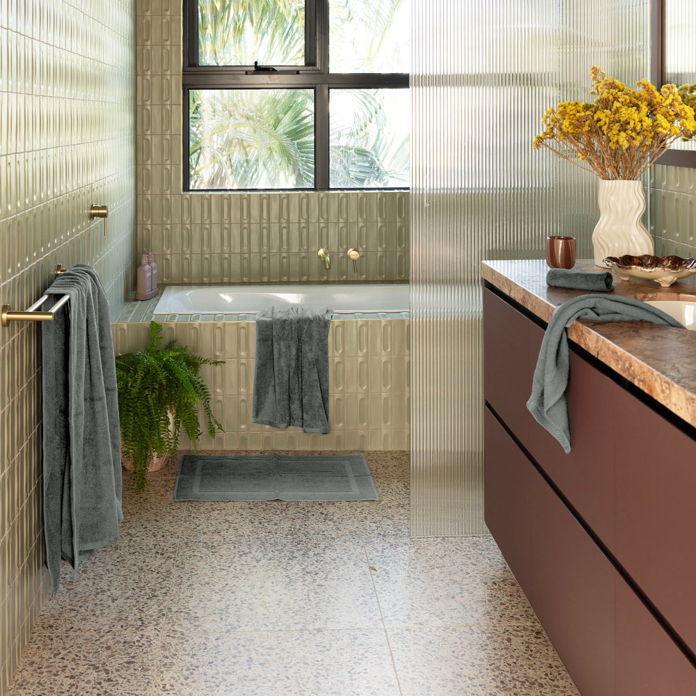 The Bambury Chateau bath mat in charcoal is placed on the floor in front of the bathtub which the rest of the charcoal collection scattered around the bathroom.