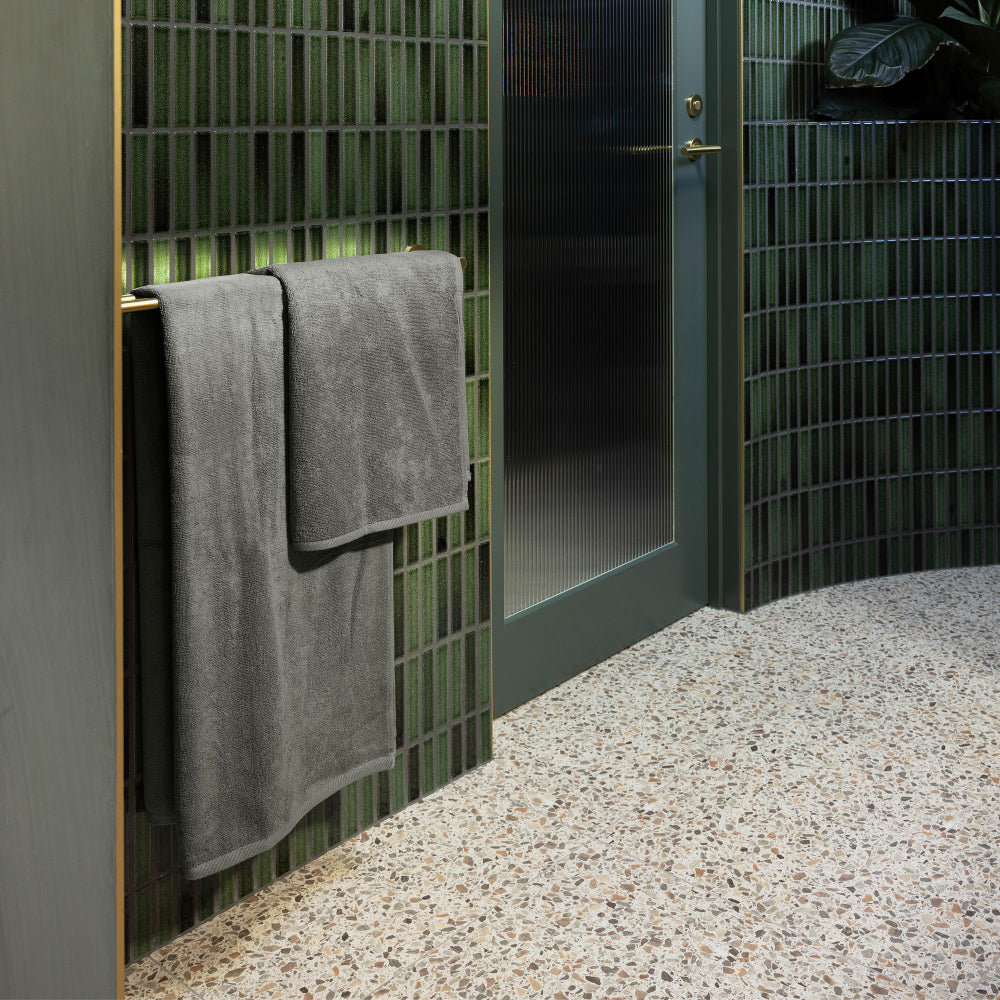 The Bambury Chateau bath mat and bath sheet are hanging on the towel rail of a bathroom featuring different shades of green complementing the charcoal of the mat and towel.