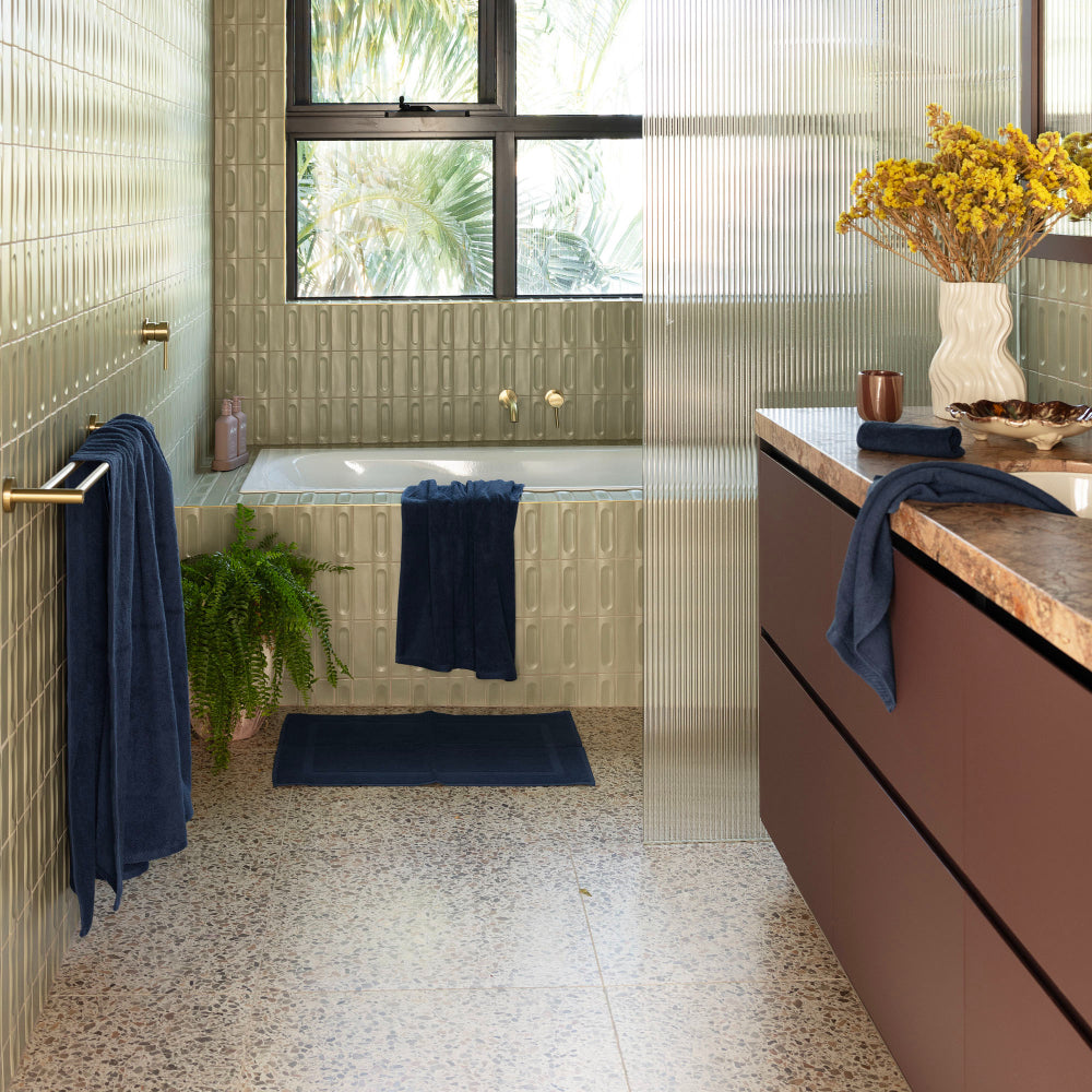 The navy Bambury Chateau bath range is being used around the bathroom, including the bath sheet on the towel rail, the bath towel hanging over the bath tub, and the bath mat layed out in front of the bath ready to enjoy a relaxing bath.